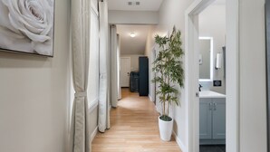 A hallway with a wooden floor, a large potted plant, a framed artwork, and a view into a room with a sink and cabinets. - 4 Queen Beds | High Speed Wifi (Fort Worth)