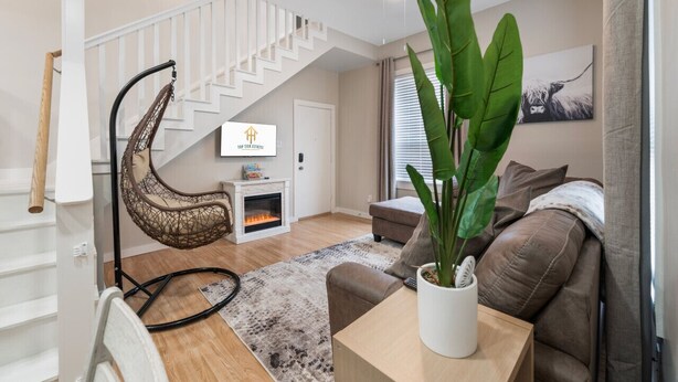 A modern living room with a white staircase, a hanging chair, a fireplace, a large plant, and a sofa. - 4 Queen Beds | High Speed Wifi (Fort Worth)
