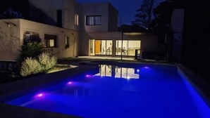 A swimming pool with blue lighting at night, surrounded by a modern house with large windows. - La Tribu, House with pool and whirlpool, ideal for families (Sierra de los Padres)