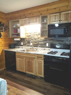 A kitchen with wooden cabinets, a stone backsplash, a stainless steel sink, a microwave, and a gas stove. - 4BR 2BA The Pine Place Cabin in Crosby - Roam, Ride, Relax  (Crosby)