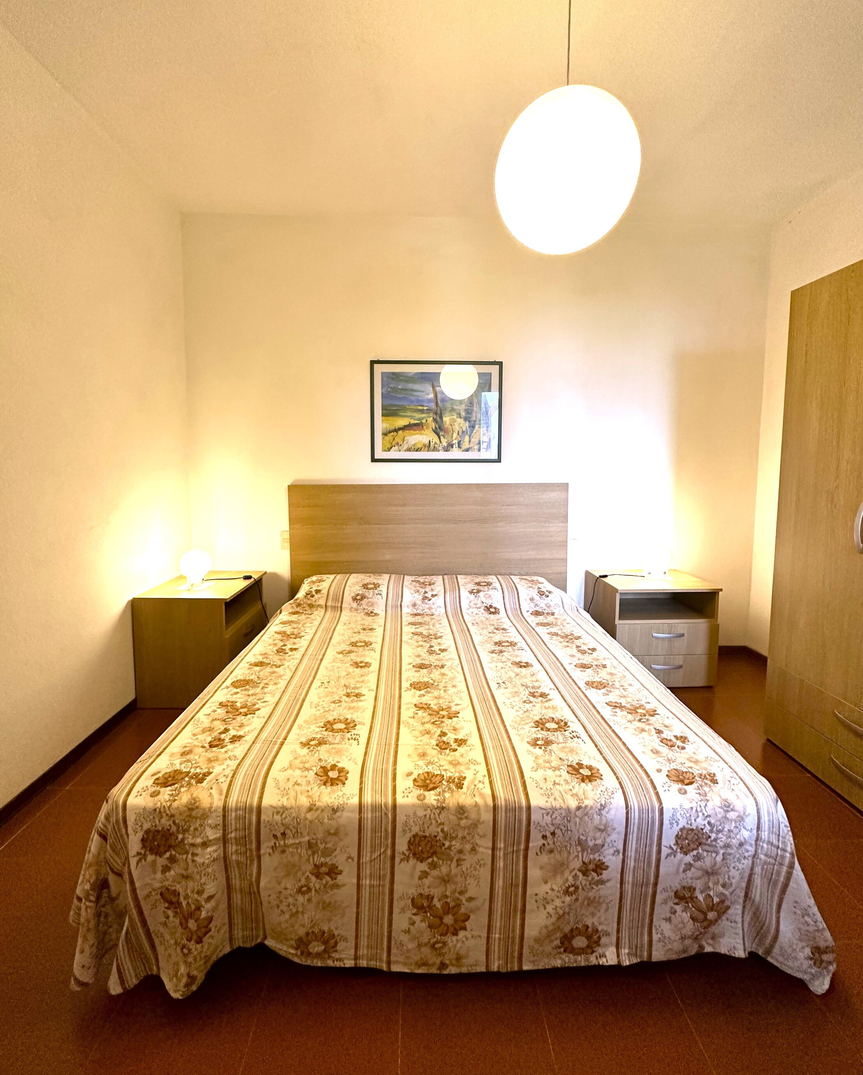 A bedroom with a bed, two bedside tables, a nightstand, a framed picture on the wall, and a wardrobe.