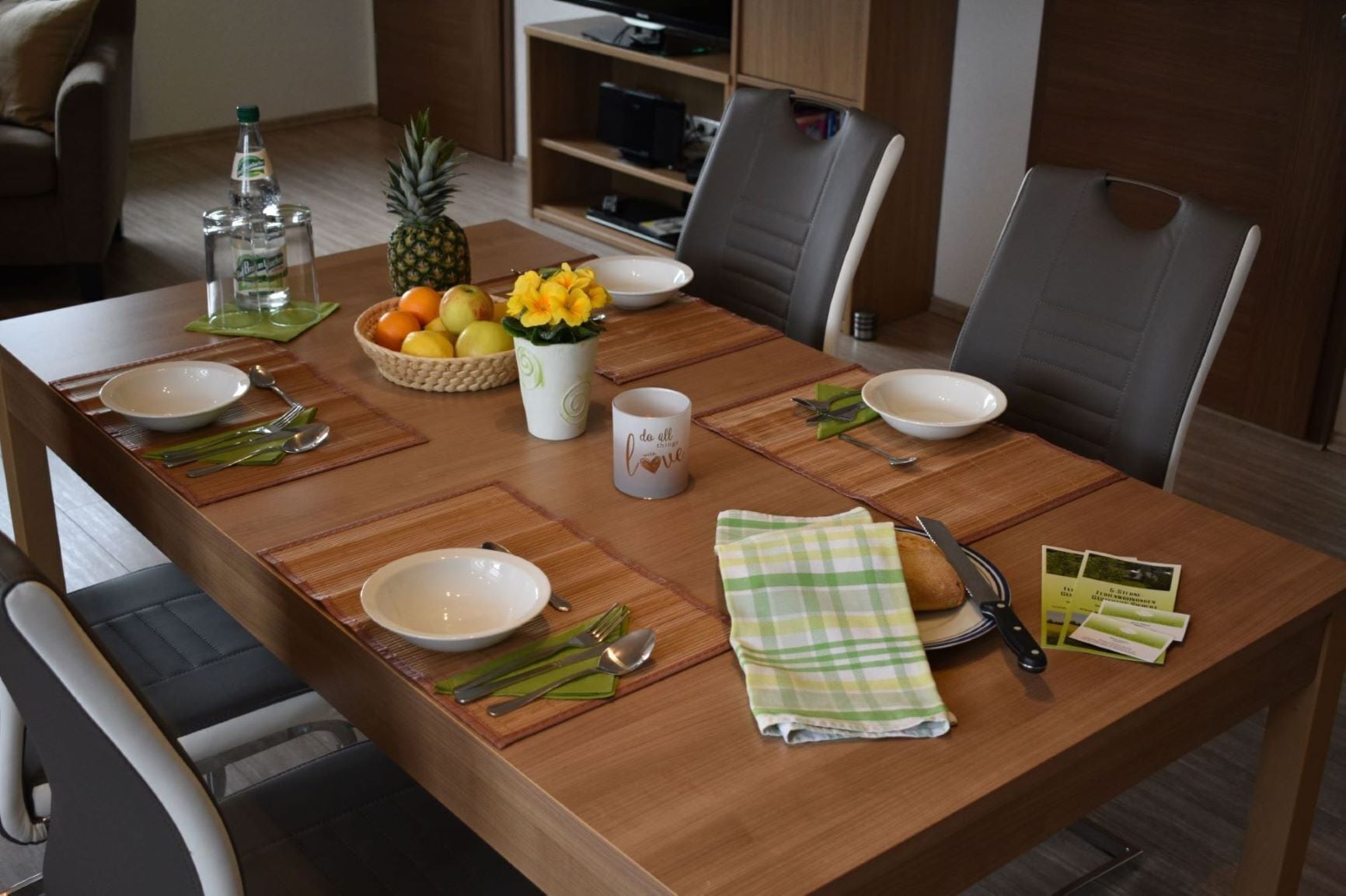 Une table à manger dressée avec des assiettes, des bols, des couverts, un ananas, un saladier de fruits, une bouteille d’eau et une tasse.