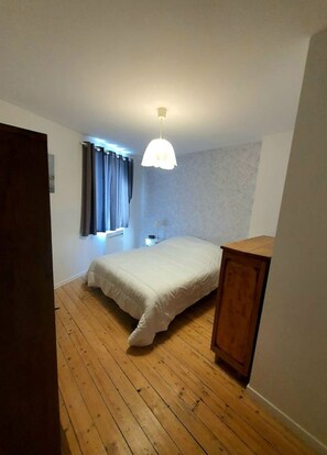 Un dormitorio con suelo de madera, cama con edredón blanco, lámpara colgante y ventana con cortinas.