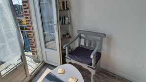Una stanza piccola con una porta scorrevole in vetro che dà su un balcone, una sedia bianca con un cuscino viola, un tavolino rotondo con due libri e un quadro raffigurante una barca al muro.