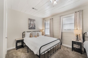 Une chambre à coucher comprenant un lit, deux tables de chevet, une lampe, une fenêtre avec des rideaux et un tableau accroché au mur.