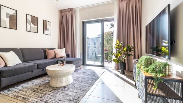 A modern living room with a sofa, coffee table, and a TV. There are curtains, a chandelier, and a balcony with plants. - Fantastic 1BR Apartment with Parking in the city center (Jerusalem)