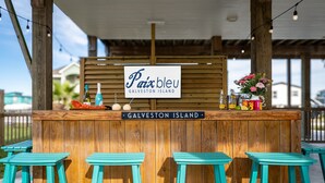 Un bar en bois avec des tabourets turquoise, une enseigne indiquant « Paix bleu, île de Galveston », et un arrangement floral.