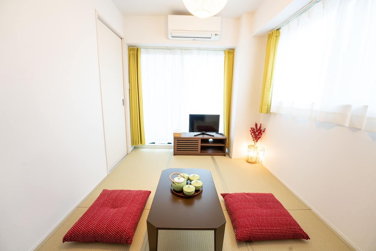 Um quarto com piso de tatame, uma mesa baixa com bandeja de chá verde, almofadas vermelhas, uma televisão e uma janela com cortinas amarelas.