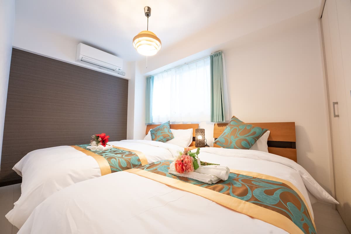 Quarto de hotel com duas camas, uma cabeceira de madeira, uma janela com cortinas, um aparelho de ar condicionado e um lustre.