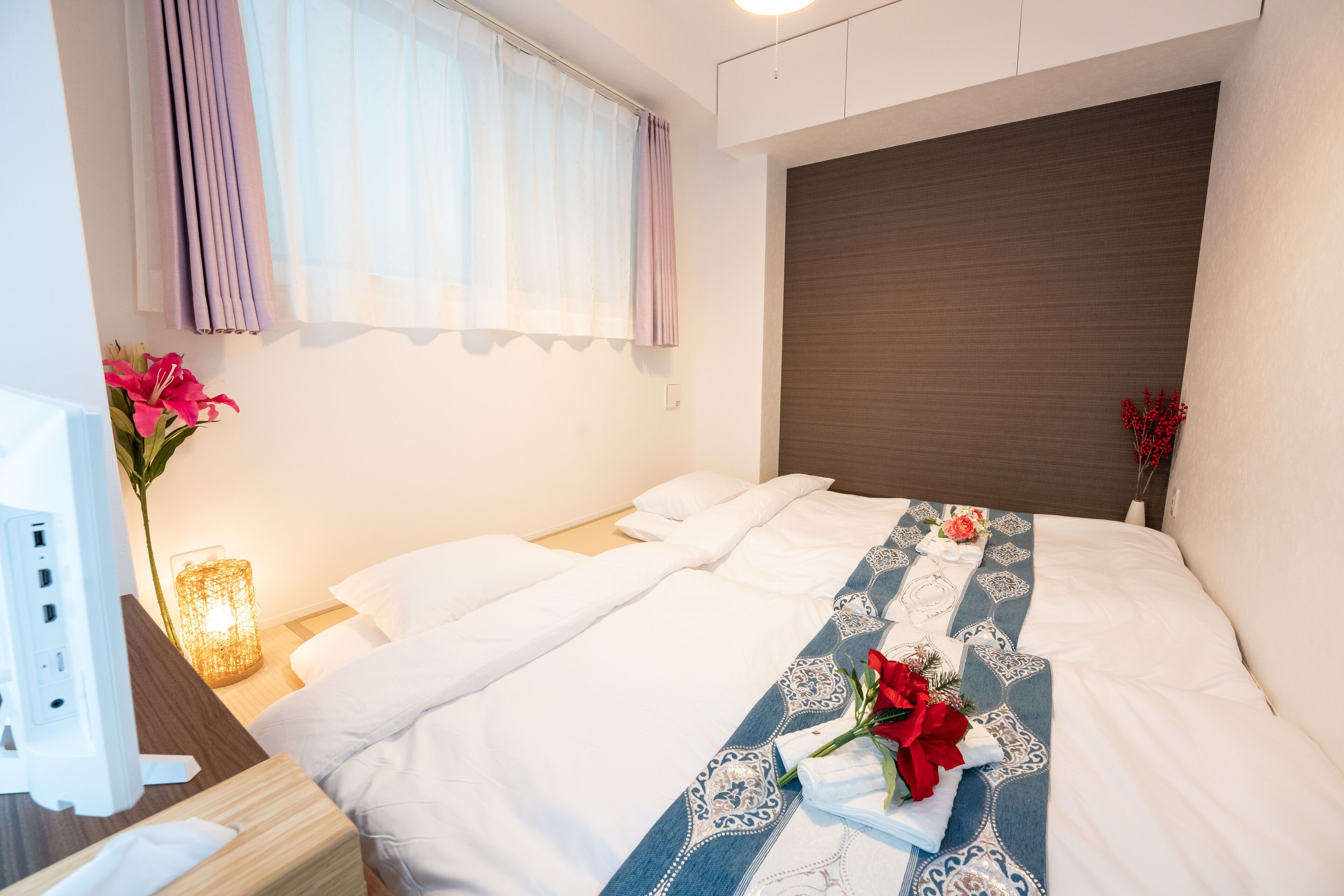 A neatly made bed with a decorative blue and white runner, a bedside lamp, and a vase with flowers.