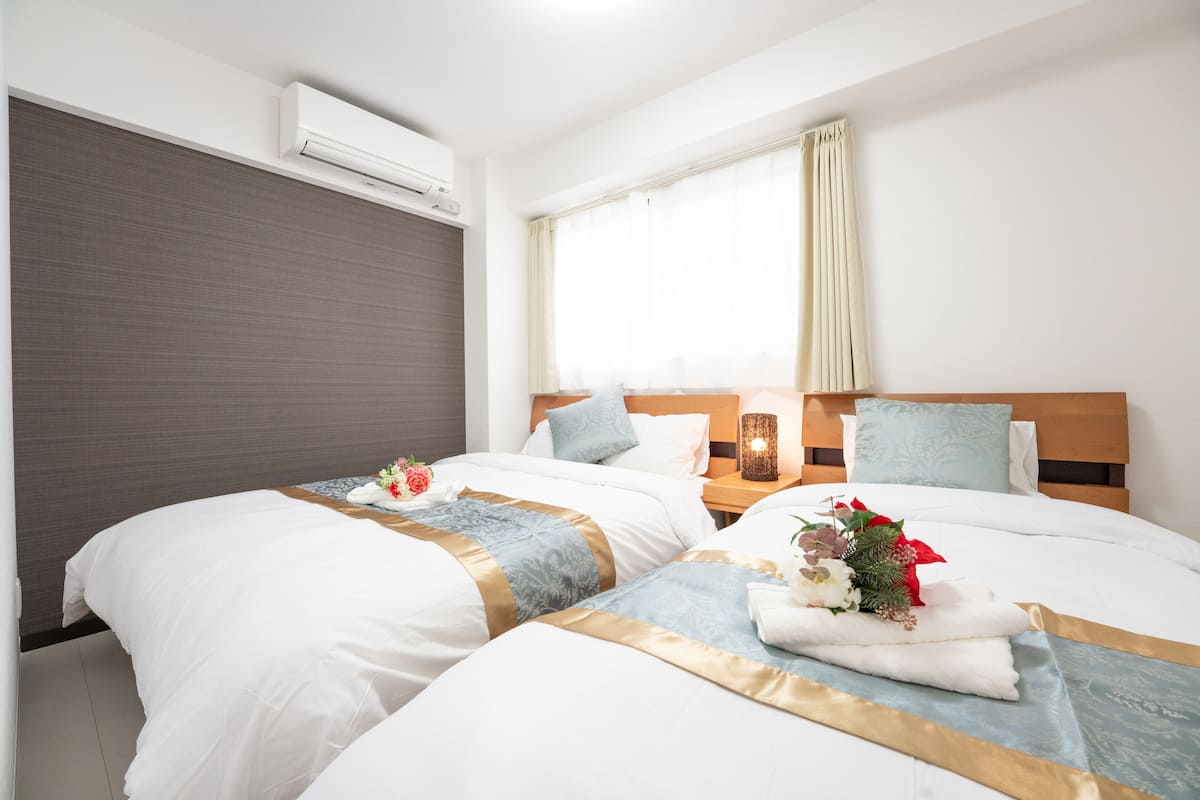 Quarto de hotel com duas camas, uma janela com cortinas, um ar condicionado e uma mesa de cabeceira com luminária.