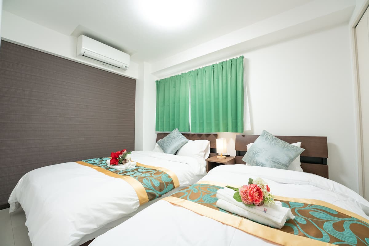 Quarto de hotel com duas camas, cortina verde, ar condicionado fixado na parede e mesa de cabeceira com abajur.