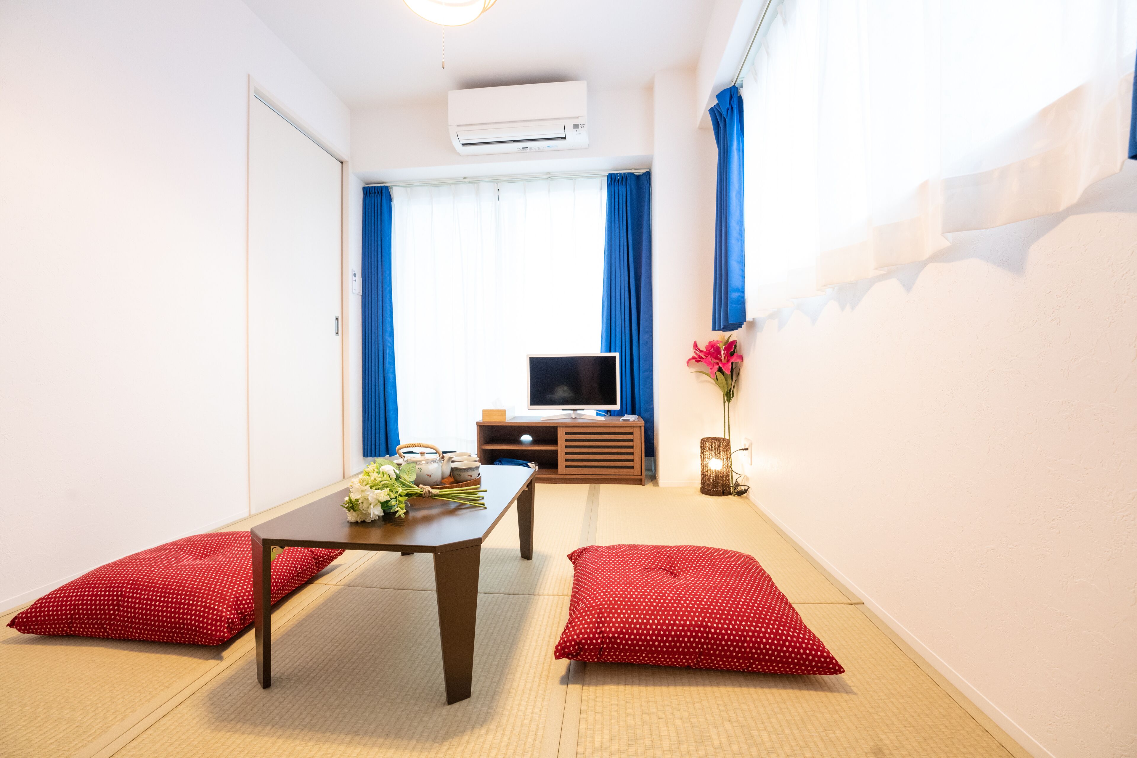 A modern hotel room with a wooden floor, a tatami mat area, a TV, a small wooden cabinet, and a red polka dot pillow.