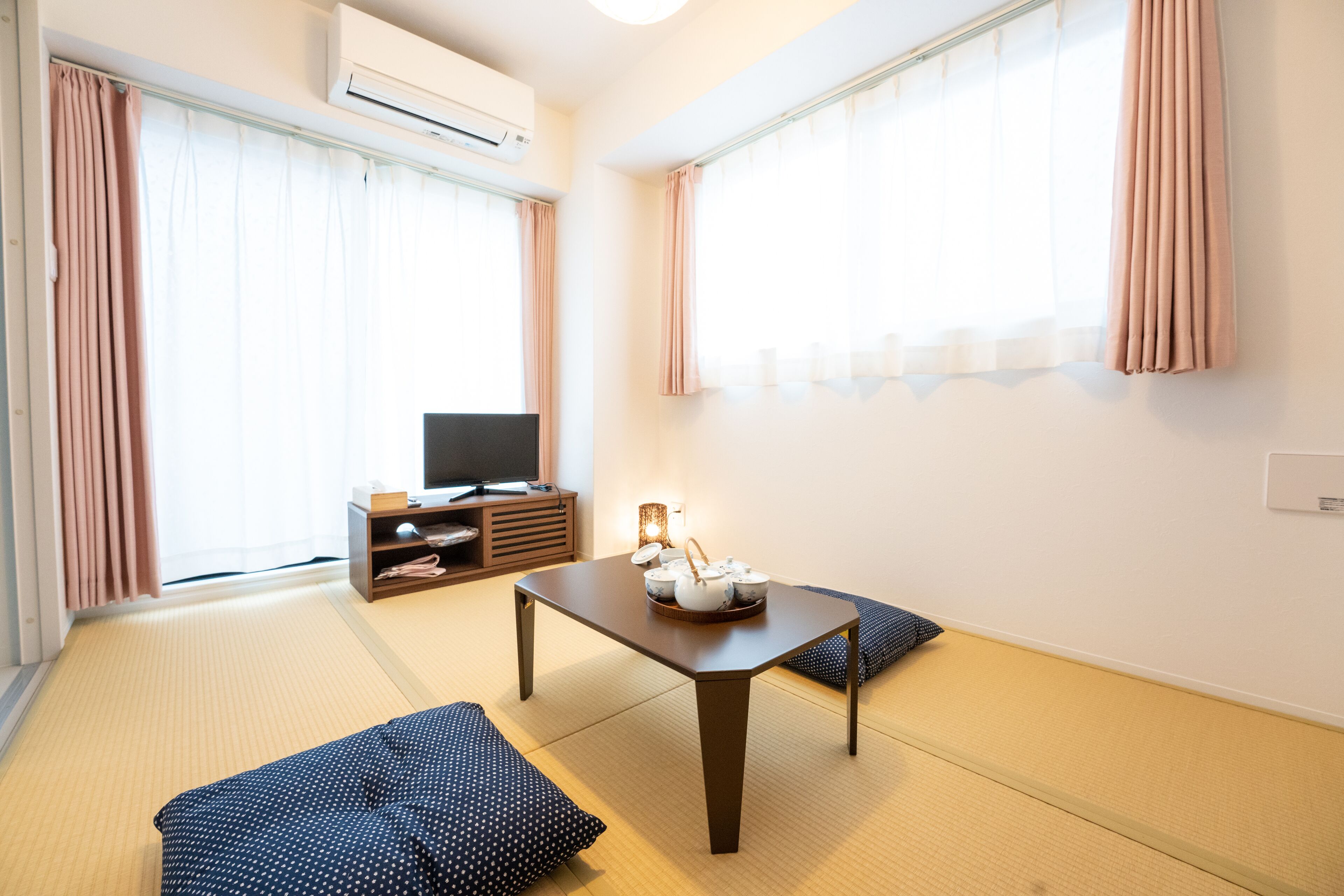 Quarto com piso de tatame, uma mesa baixa com um bule, televisão em um móvel e uma porta deslizante com cortinas.