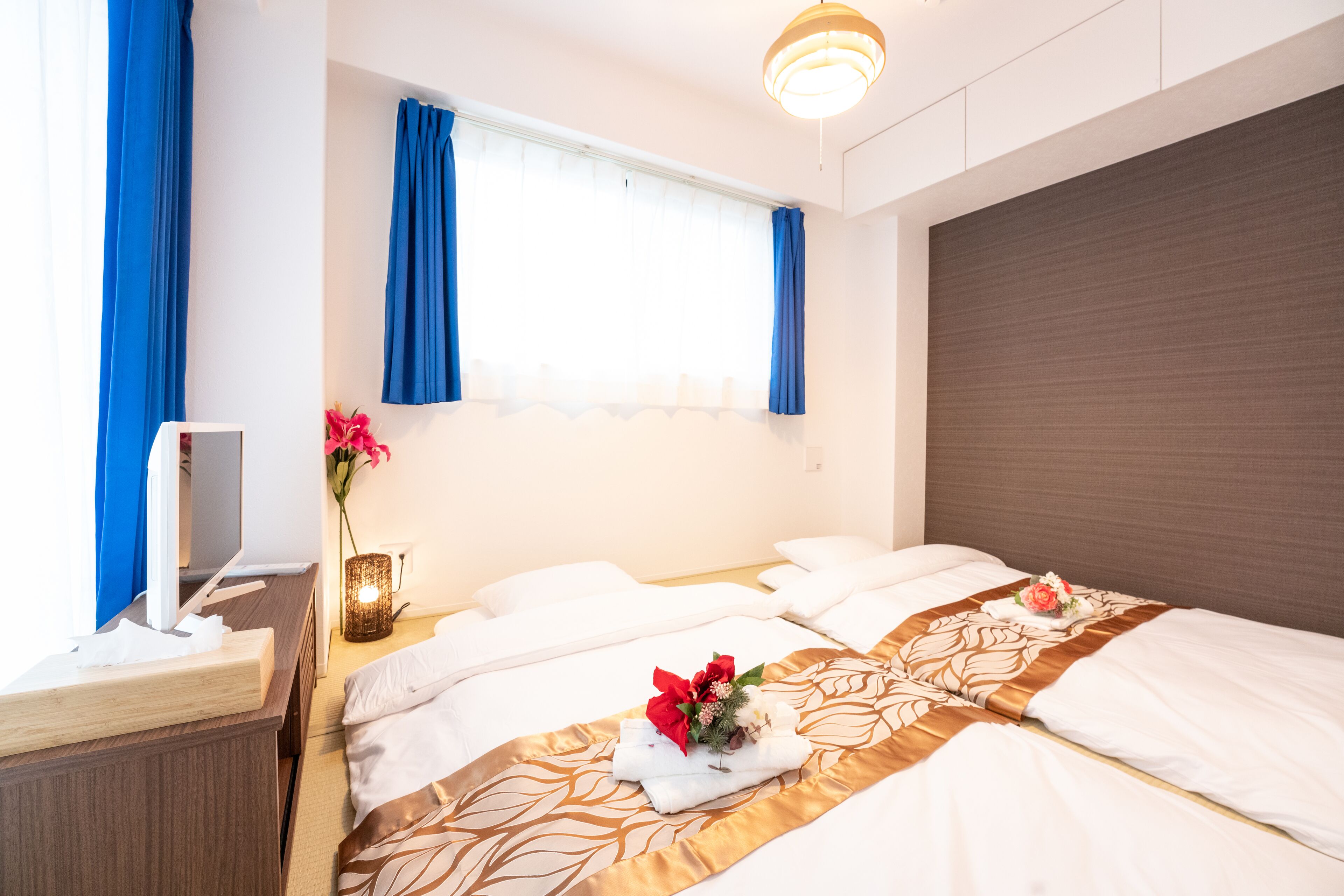 A hotel room with two beds, a desk with a computer, a vase with flowers, and a window with curtains.