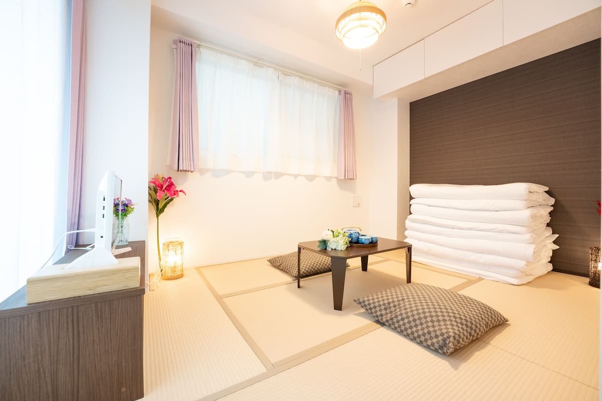 Quarto de hotel com piso de tatame, cama com lençóis brancos, mesa de cabeceira com um vaso de flores e janela com cortinas leves.