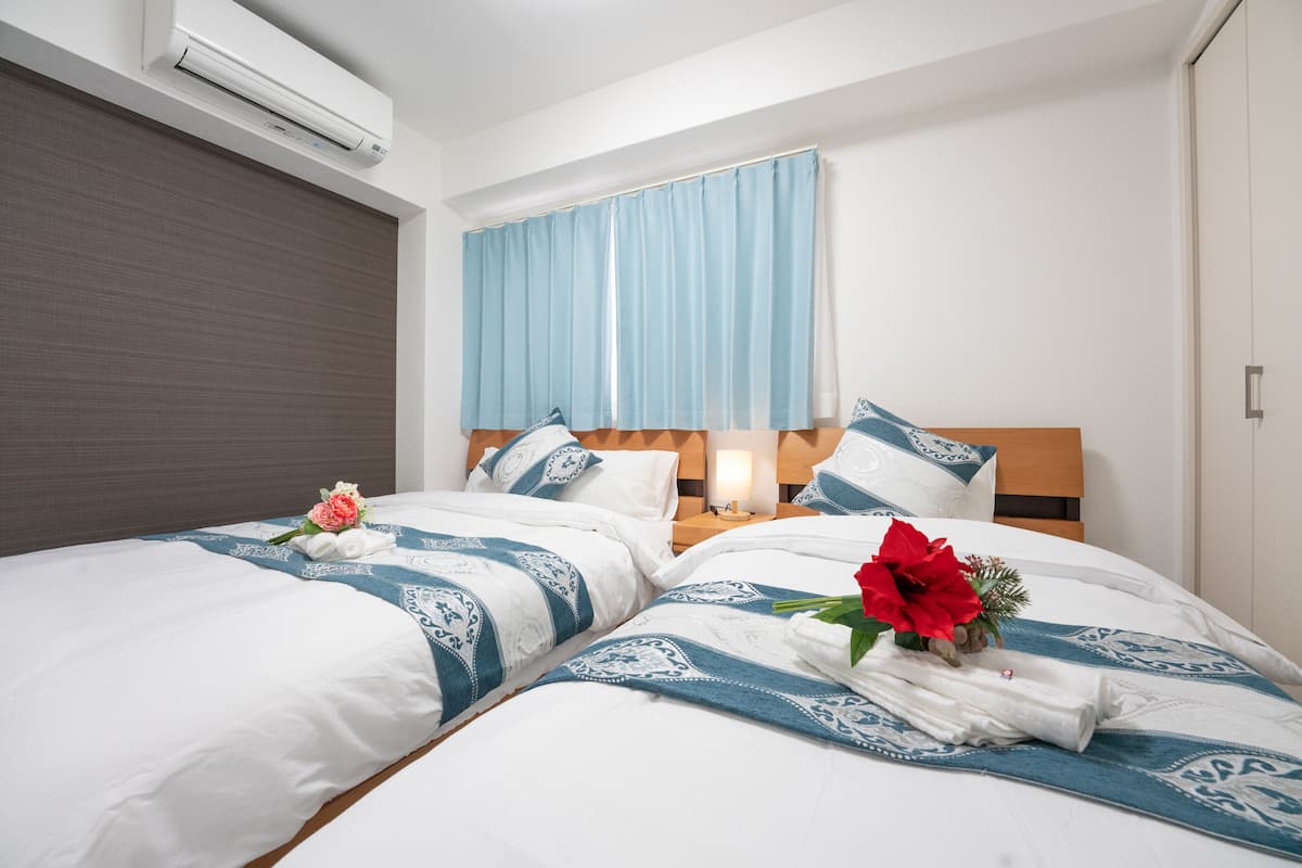 Quarto de hotel com duas camas, uma janela grande com cortina azul, ar condicionado e cabeceira de madeira.