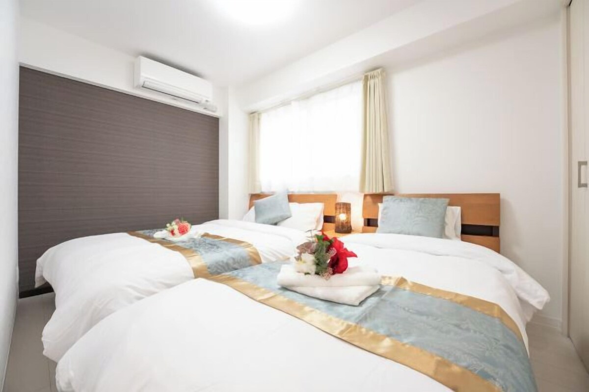 Quarto de hotel com duas camas, uma janela grande com cortinas, aparelho de ar condicionado e mesa de cabeceira com abajur.