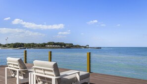 A deck with two white lounge chairs facing a calm sea, a clear sky, and a distant island with buildings.