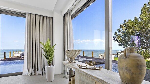 Ein modernes Zimmer mit großem Fenster mit Blick auf einen Pool und den Ozean. Es gibt einen Topfpflanzen, eine dekorative Vase und einen Marmortisch mit Tablett.
