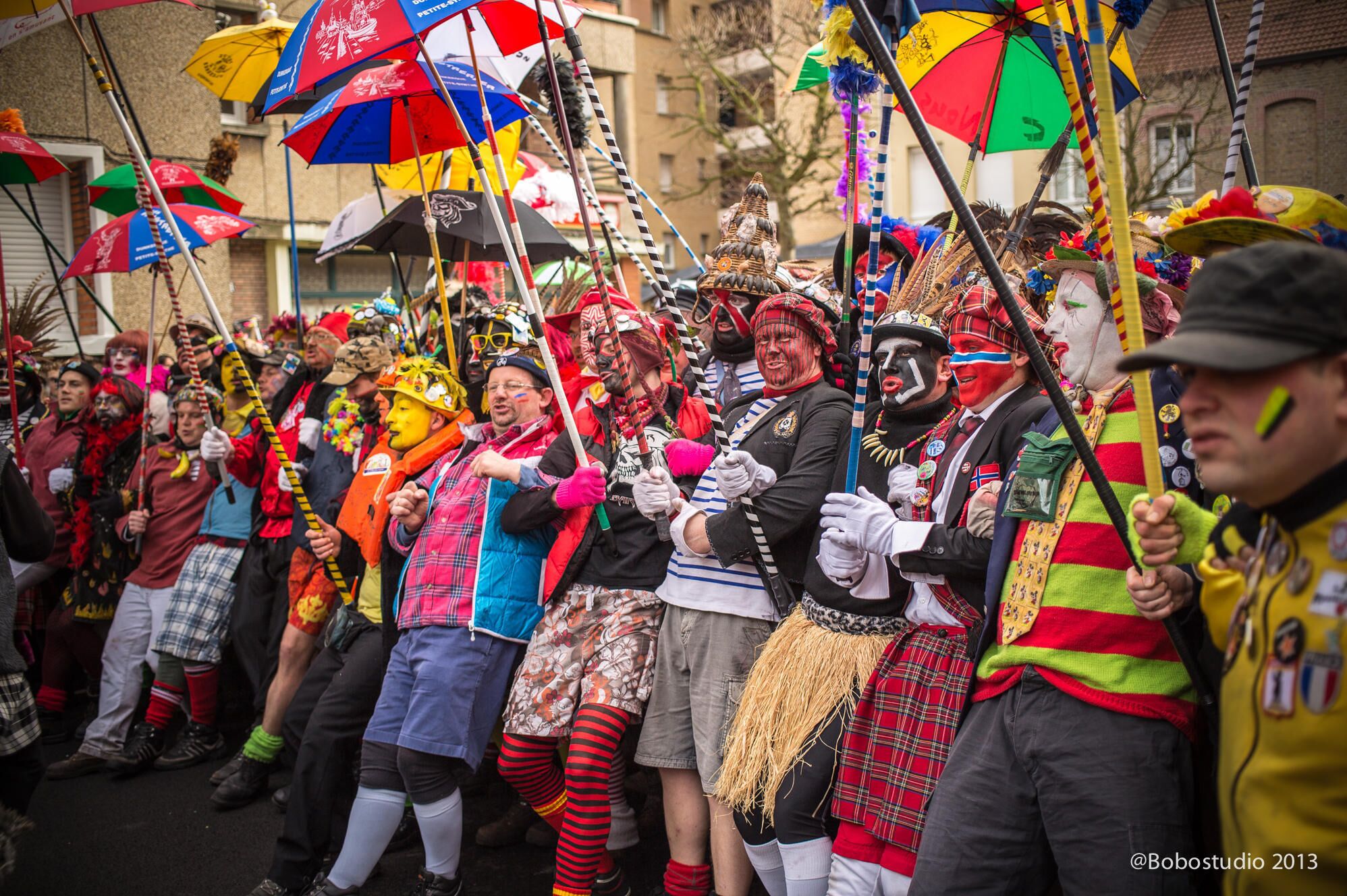 Un grupo de personas con trajes y máscaras coloridos, llevando paraguas y banderas, participan en un desfile o festival.