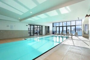 A large indoor swimming pool with clear blue water, surrounded by sunlit rooms and large windows offering views of the city.