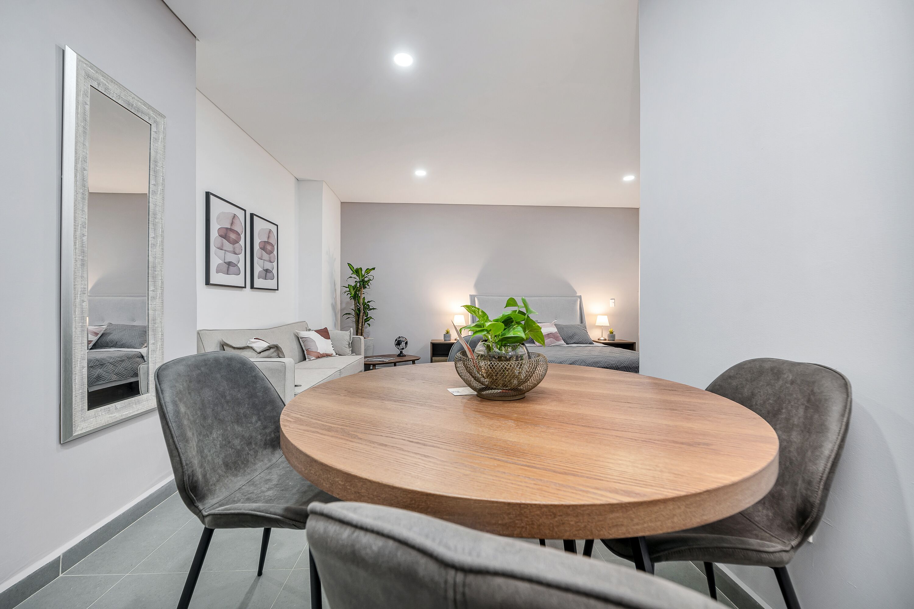 A modern dining area with a round wooden table, two grey upholstered chairs, a decorative plant in a woven basket, and a large mirror on the wall.