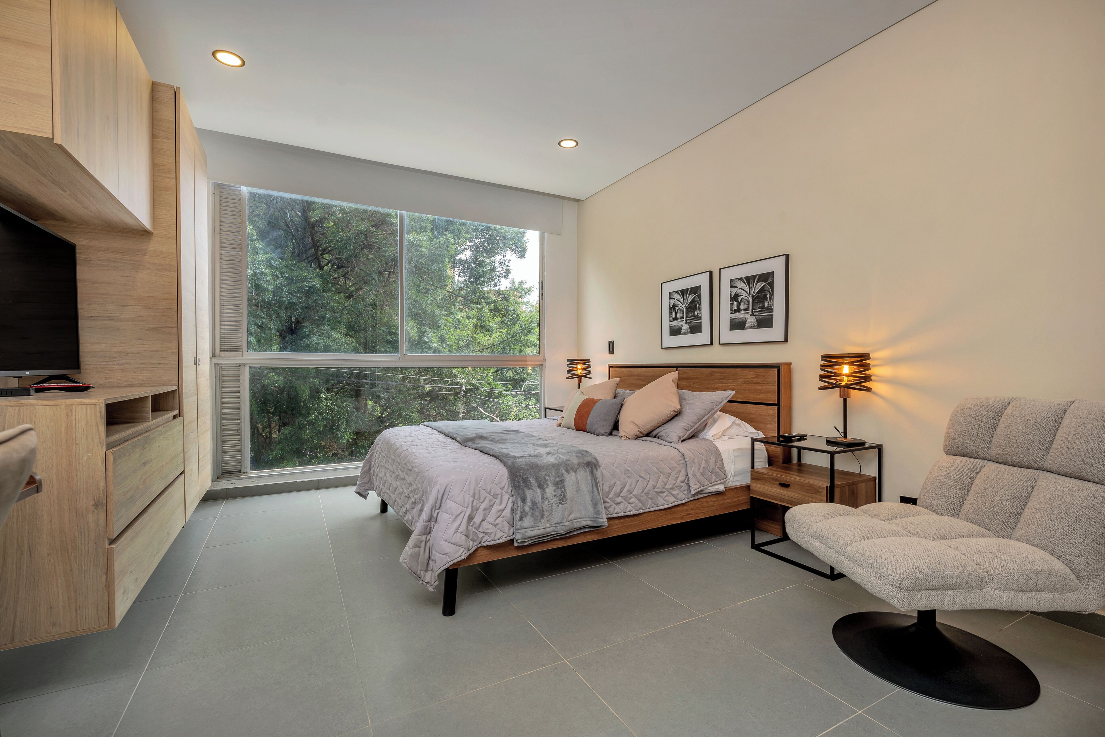 Un dormitorio moderno con un ventanal grande, una cama con colcha gris, una mesita de noche de madera, un sillón gris y un televisor sobre un mueble de madera.