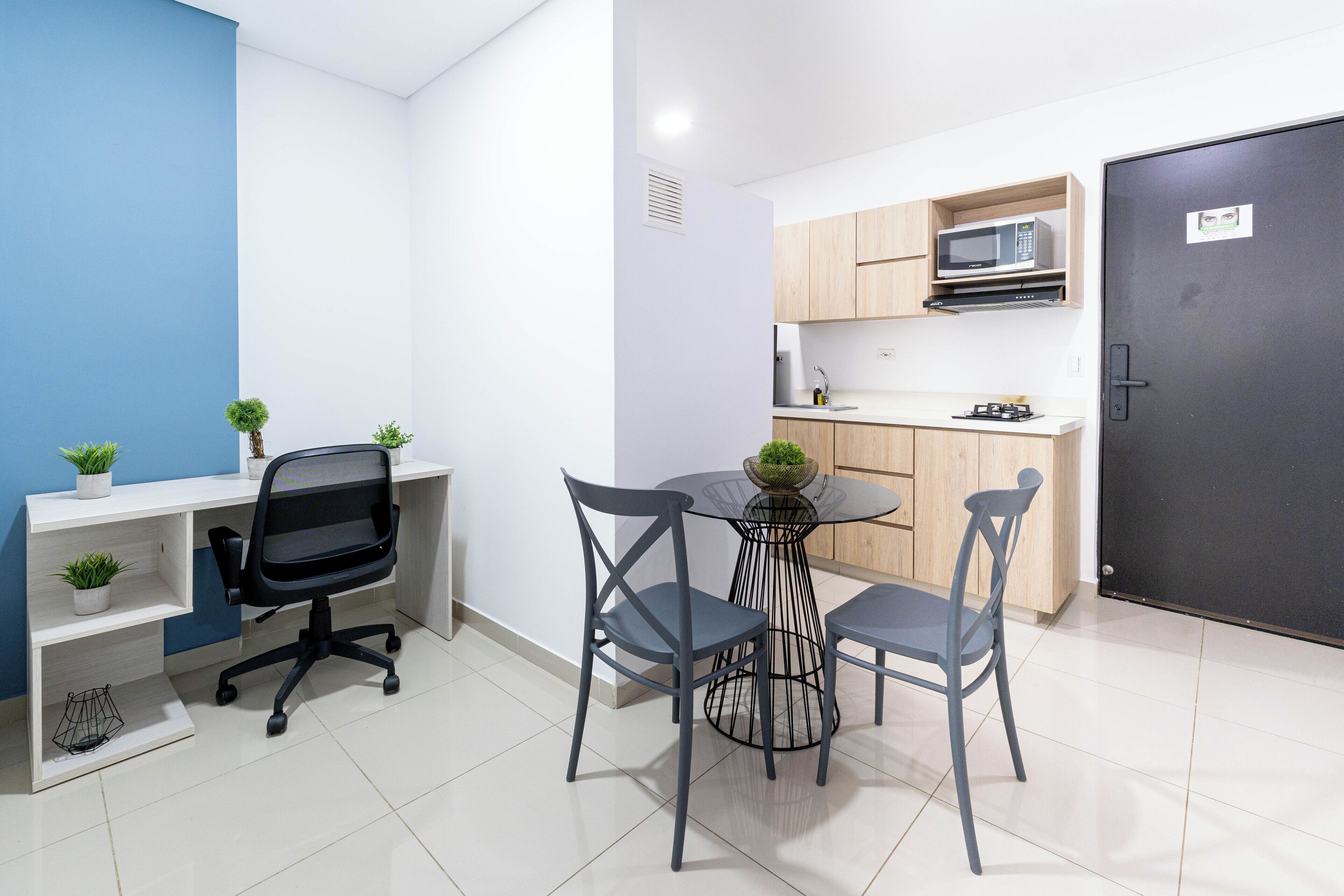 A compact kitchen with a microwave, a small sink, and wooden cabinets. There is a dining table with four chairs and a potted plant on it.