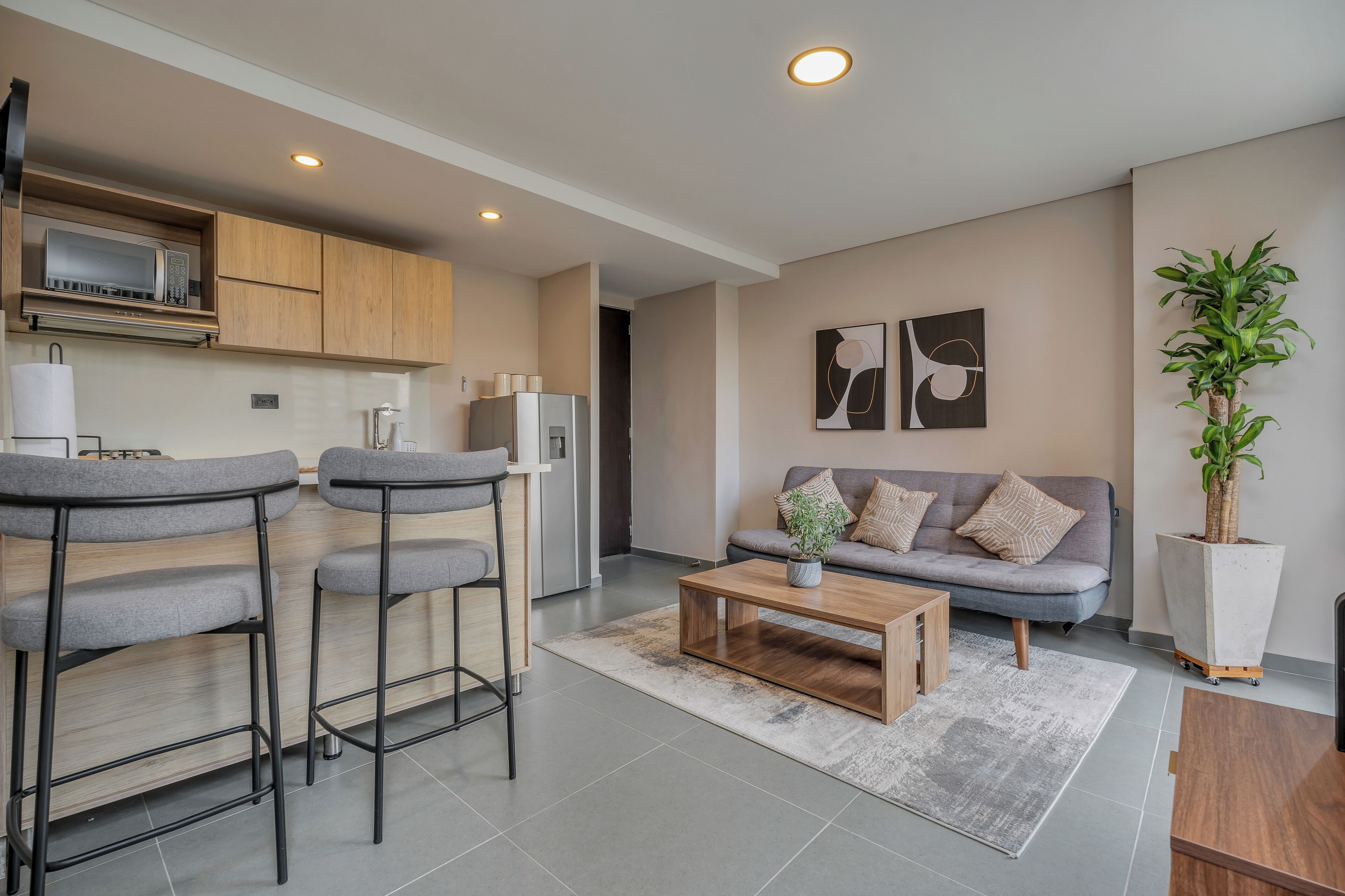 A modern kitchen with a microwave, refrigerator, and bar stools. There is a grey sofa with cushions and a coffee table in the living area.