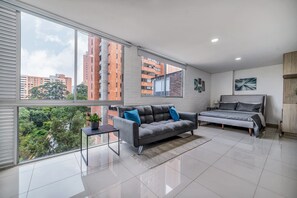 Ein modernes Wohnzimmer mit grauem Sofa, einem Bett und einem großen Fenster mit Blick auf die Stadt.