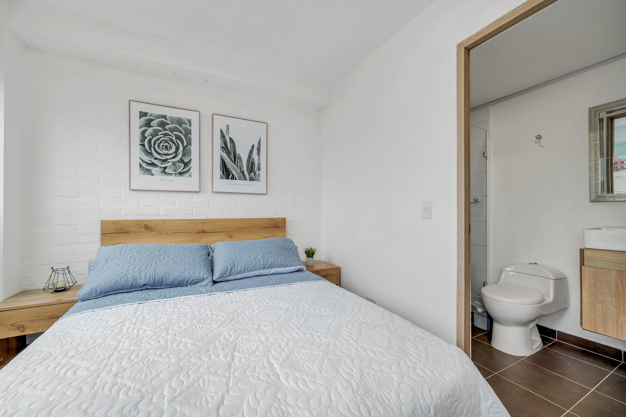 Un dormitorio con cama, mesita de noche, una planta pequeña y un baño con inodoro y lavamanos.