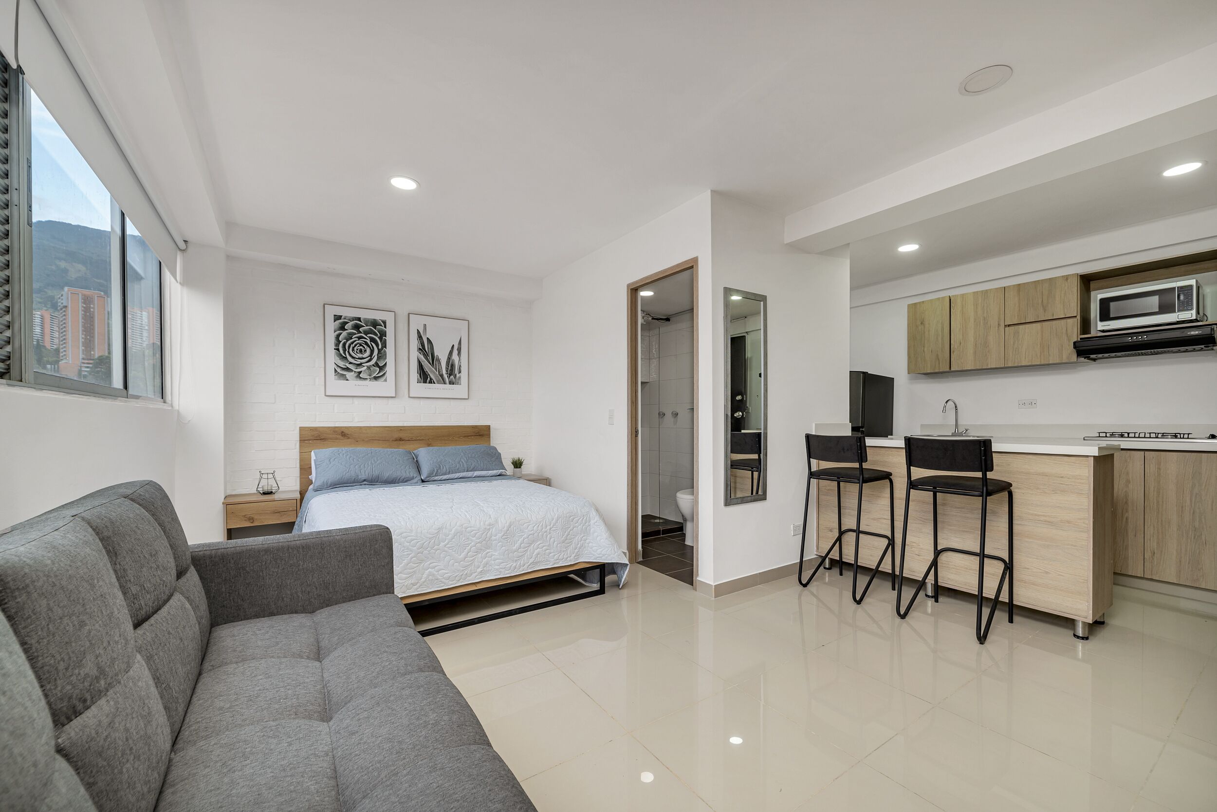A modern bedroom with a bed, a grey sofa, and a small bedside table. There is a kitchen area with a microwave, a sink, and bar stools.