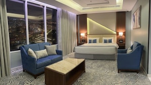 Habitación de hotel con una cama grande, un sofá, una silla y vistas a un paisaje urbano a través de amplios ventanales.
