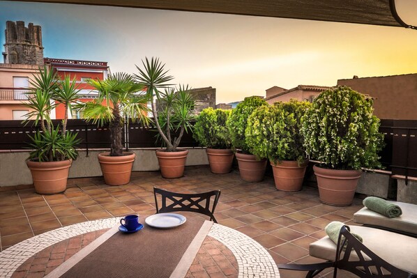 A rooftop terrace with potted plants, a table with chairs, and a view of buildings in the background.
