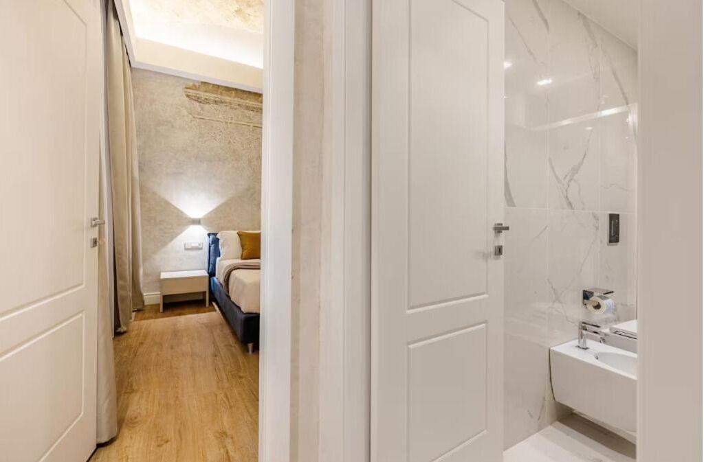 A hotel room with a wooden floor, a bed with a white headboard, a bedside table, a lamp, and a bathroom with a white sink and mirror.