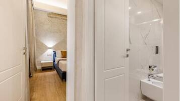 A hotel room with a wooden floor, a bed with a white headboard, a bedside table, a lamp, and a bathroom with a white sink and mirror.