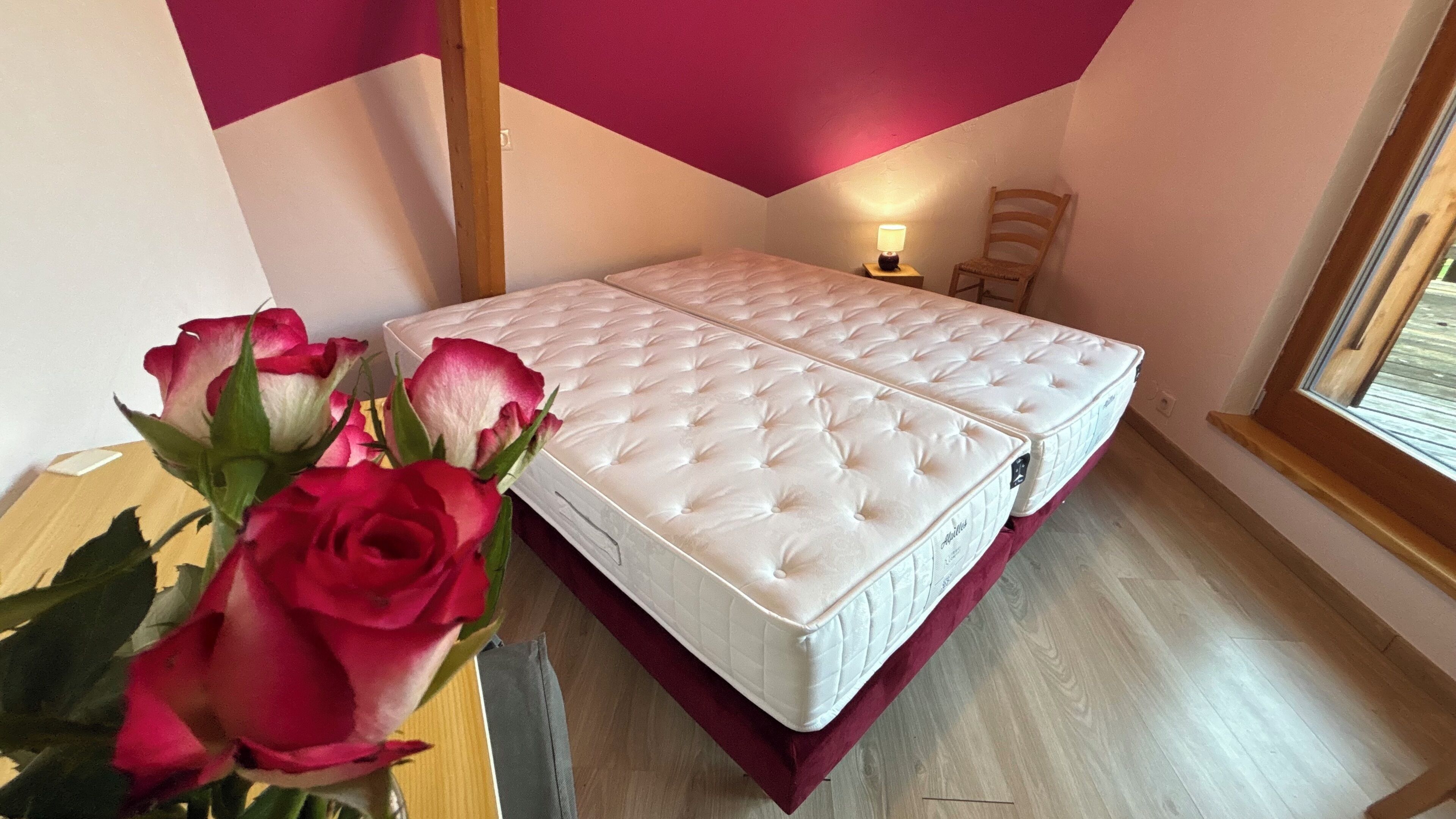 Ein Schlafzimmer mit zwei weißen, gepolsterten Matratzen, einem Holzstuhl, einem Nachttisch mit Lampe und einem Fenster mit Blick.