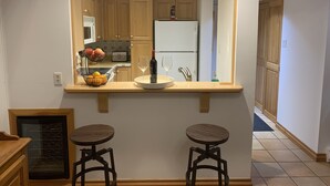 A kitchen with wooden cabinets, a built-in wine cooler, a fruit basket, and two bar stools.