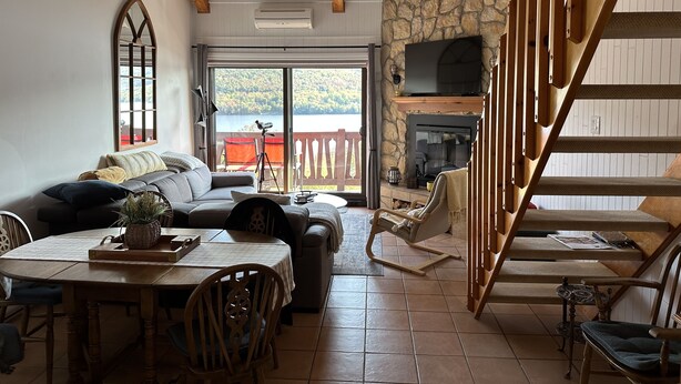 A cozy living room with a stone fireplace, wooden staircase, and a large window overlooking a scenic view.