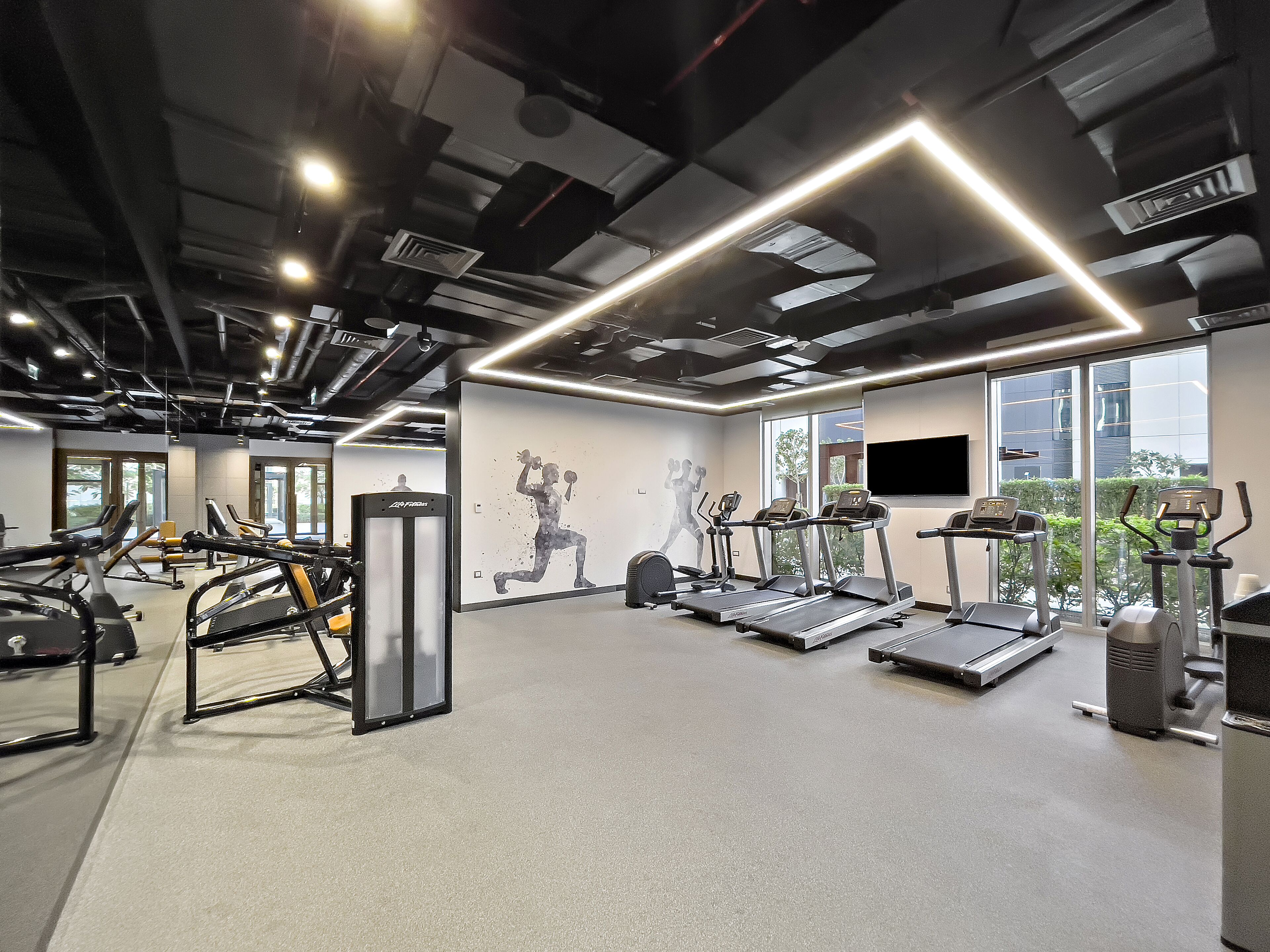 Une salle de sport moderne équipée de tapis de course, d’appareils de musculation et dotée d’une grande fenêtre offrant une vue sur la ville.