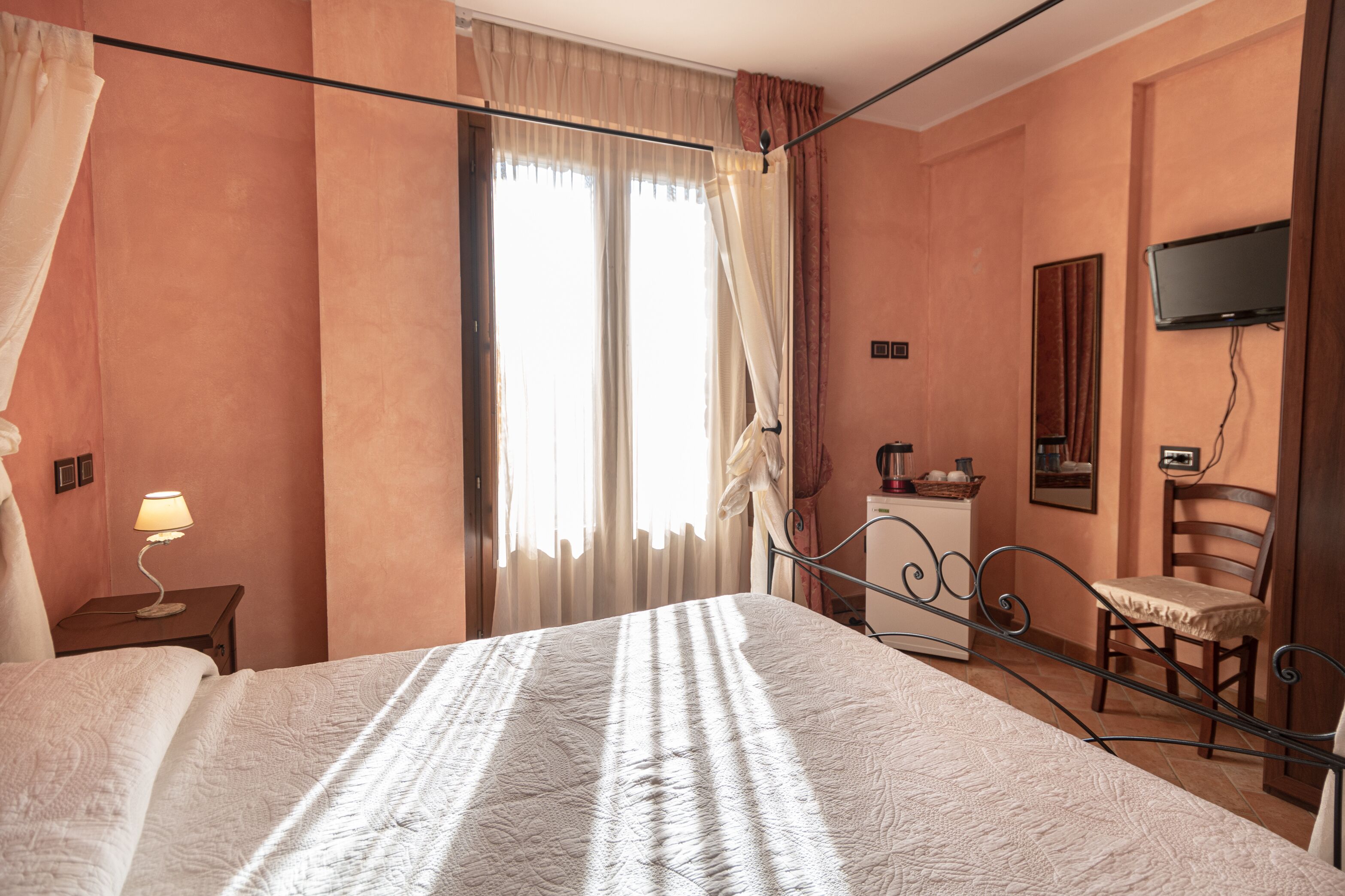 Ein Schlafzimmer mit einem Bett, einem Nachttisch mit Lampe, einem Sessel, einem an der Wand befestigten Fernseher und einem Fenster mit Vorhängen.