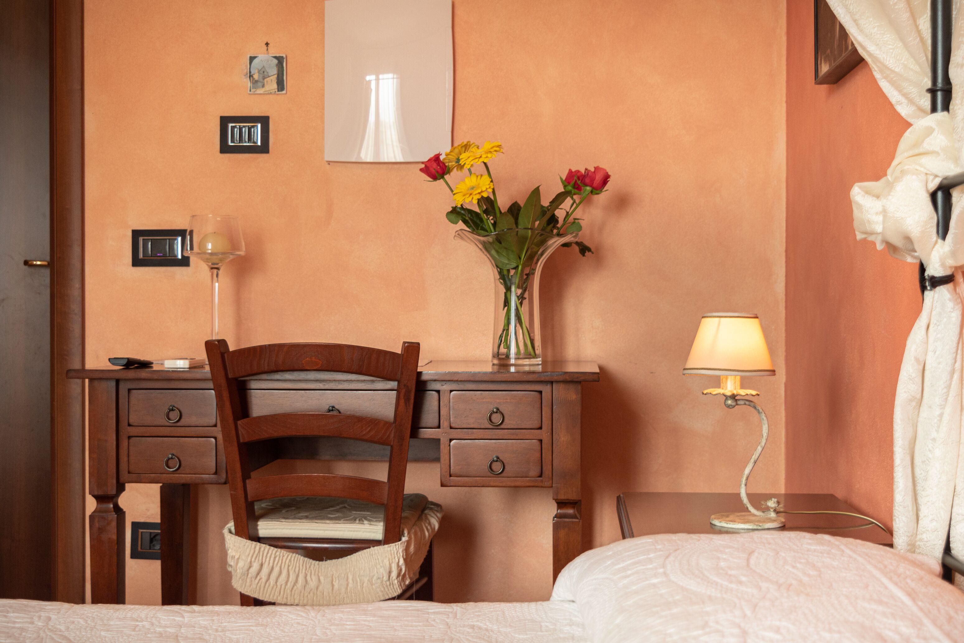 A hotel room with a wooden desk, a chair, a bed, a nightstand with a lamp, and a vase with flowers.