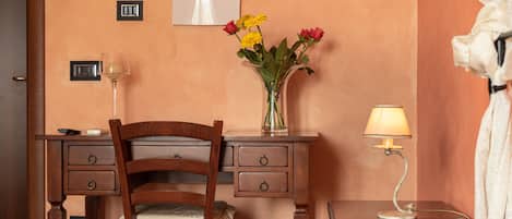 A hotel room with a wooden desk, a chair, a bed, a nightstand with a lamp, and a vase with flowers.