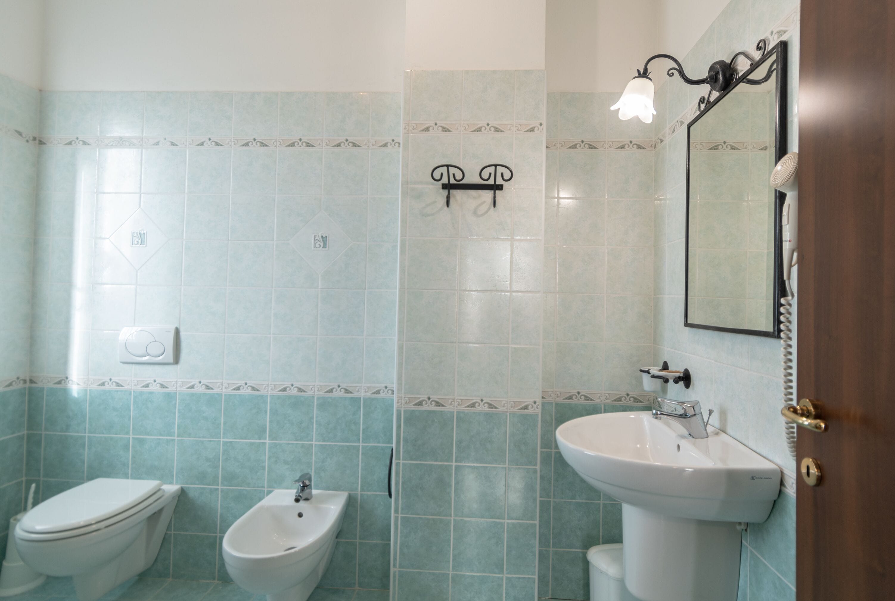 Une salle de bain équipée de toilettes, d’un bidet et d’un lavabo, dont le carrelage est de couleur bleu clair.