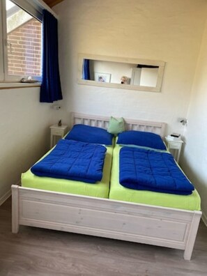 A double bed with blue bedding, a small bedside table, a nightstand, a window with curtains, and a mirror above the bed.