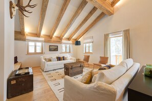 Un amplio salón con vigas de madera, un sofá blanco, una mesa de centro y un gran ventanal que proporciona luz natural.
