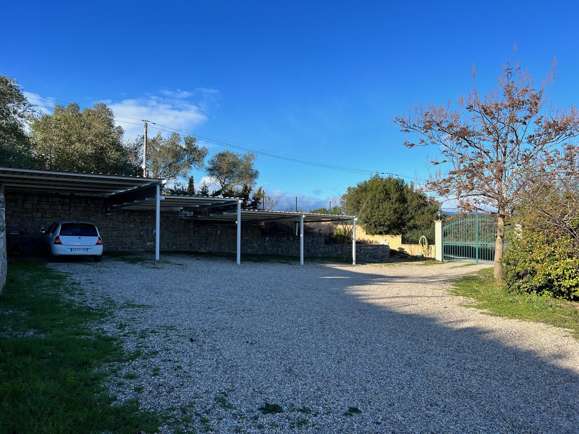 Uma entrada de cascalho leva a uma área de estacionamento coberta, com um muro de pedra e um carro estacionado lá dentro.