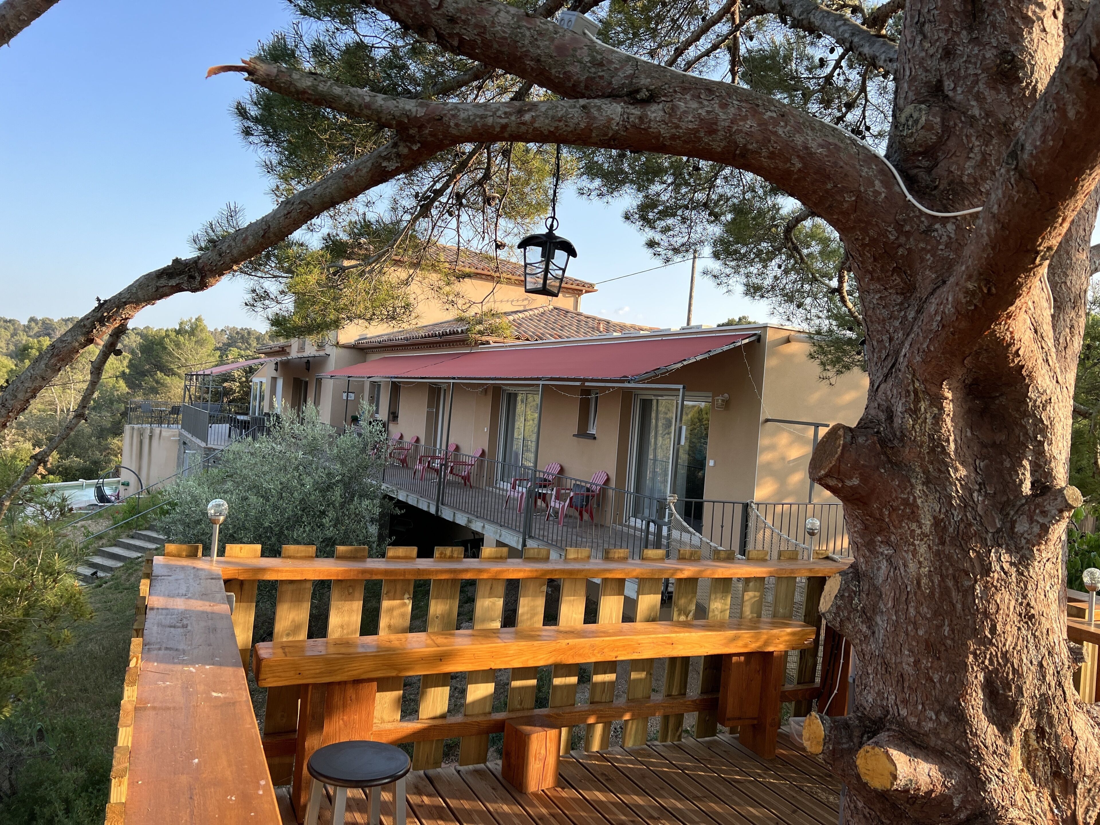 A wooden deck with a bench and a small stool, overlooking a building with a red awning and a balcony with red chairs.
