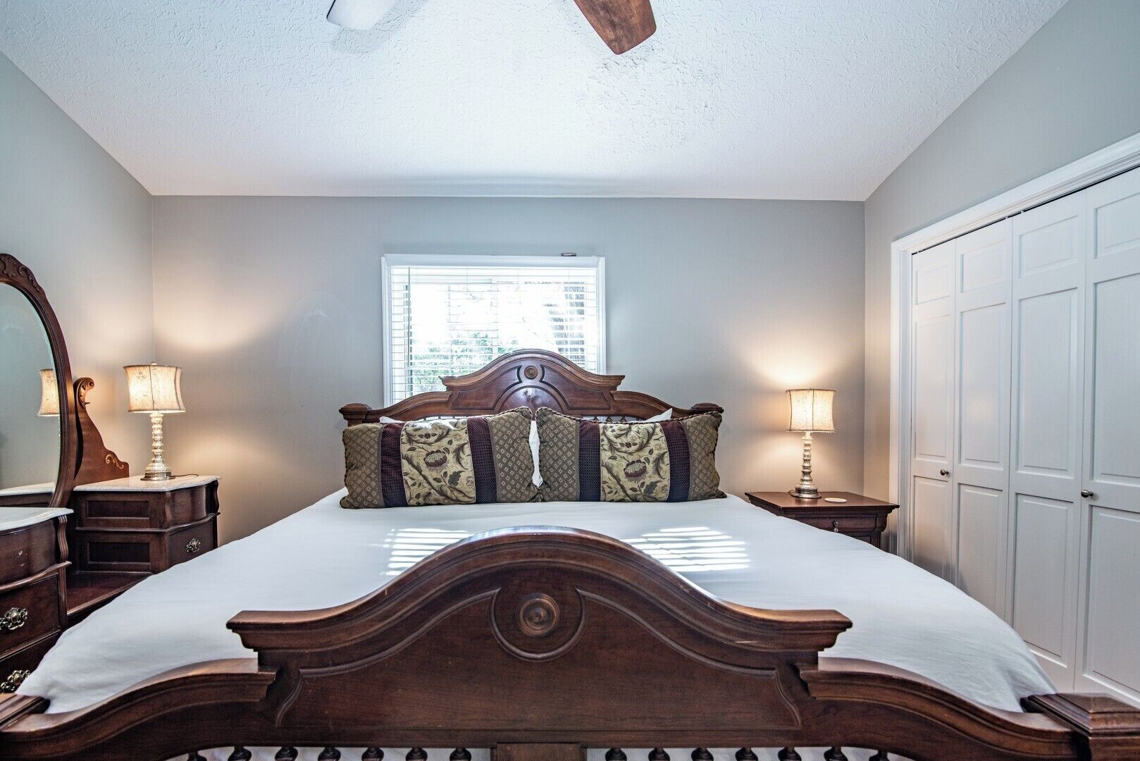 A bedroom with a large bed, wooden headboard, two bedside lamps, a mirror, and a window.
